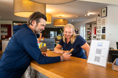 Golfer checkt an der Golfrezeption ein und fragt Rezeptionistin etwas mit Blick auf sein Handy